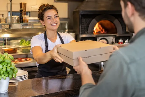 Mitarbeiterin reicht einem Kunden zwei Pizzakartons über die Theke